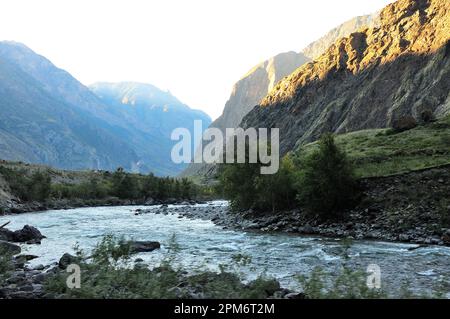 Der Schatten der untergehenden Sonne bedeckt ein wunderschönes Tal in den Bergen mit einem stürmischen Fluss, der seinen Grund entlang fließt. Chulyshman River, Alta Stockfoto