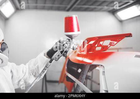 Spritzlackierer in Schutzhandschuh mit Airbrush Pulverizer, roter Autostoßfänger in weißer Lackierkammer. Stockfoto