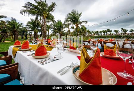 Mehrere lange Esstische draußen in einer tropischen Umgebung mit vielen Palmen und roten Servietten mit Besteck. Stockfoto