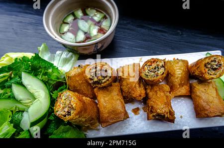 Vietnamesische Frühlingsrollen, aufgeschlitzt auf einem langen weißen Teller mit einer Seite Grüns, Gurke, Salat, Dip. Stockfoto