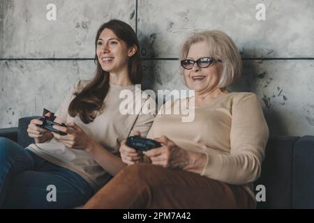 Zwei weiße, glückliche Frauen, Erwachsene Tochter und Großmutter, die mit dem Joystick Videospielkonsole spielen, während sie zu Hause mit echten Menschen in ihrer Freizeit sitzen Stockfoto