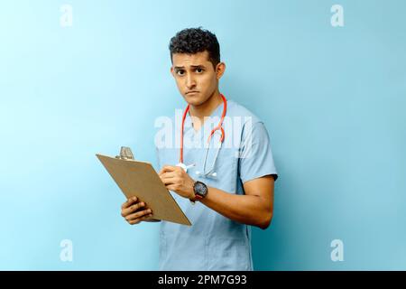 Ein junger Arzt, der eine medizinische Uniform und ein Stethoskop trägt, steht mit einem Klemmbrett in der Hand Stockfoto