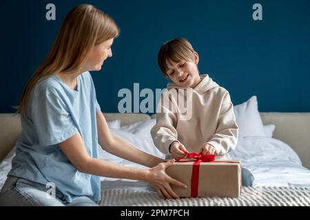 Glücklicher kleiner Junge Kind öffnet eine Geschenkbox, während er seinen Geburtstag mit seiner Mutter zu Hause feiert Stockfoto