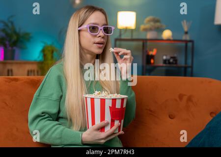 Aufgeregte Frau mit 3D-Brille sitzt auf dem Sofa und isst Popcorn-Snacks, sieht sich ein Interessantes Sportspiel an und filmt in der Wohnung Filme in sozialen Medien. Ein Mädchen, das Unterhaltung genießt Stockfoto