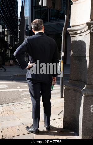 Rückansicht eines Geschäftsmanns, der am 4. April 2023 in London, England, an einer Straßenecke in der City of London, auch bekannt als The Square Mile, im Finanzviertel der Hauptstadt, allein steht. Stockfoto