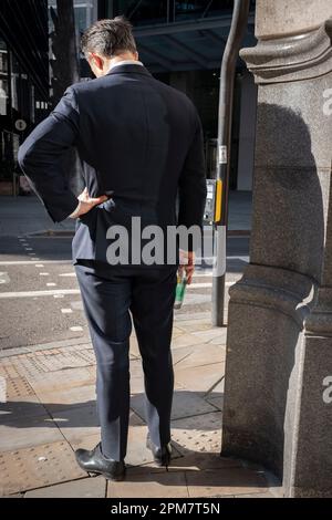 Ein Rückblick auf einen allein stehenden Geschäftsmann mit Blick auf den Boden an einer Straßenecke in der City of London, auch bekannt als die Square Mile - das Finanzviertel der Hauptstadt, am 4. April 2023 in London, England. Stockfoto