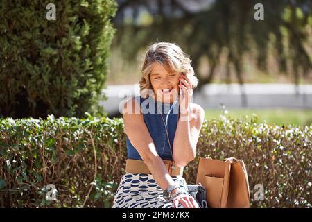 Ältere Frau, die telefoniert, während sie auf einer Bank sitzt: Eine reife Frau in ihren 60s Jahren sitzt bequem auf einer Parkbank, während sie mit ihrem Handy spricht. Stockfoto