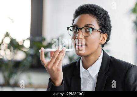 Lächelnde afroamerikanische Geschäftsfrau in schwarzem Blazer und Brille, die Sprachnachricht auf Smartphone im Büro aufnimmt, Stock Image Stockfoto