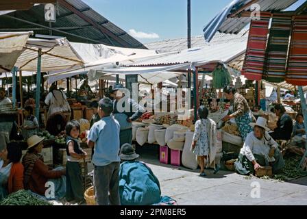 Ein geschäftiger Markt für Erzeugnisse unter freiem Himmel in Cochabamba, Bolivien im Jahr 1968. Obst- und Gemüsehändler wetteifern unter Welleisendächern auf ihren Verkaufsständen um den Handel. Ein Schild weist darauf hin, dass auch Öl (Aceite) verkauft wird. Cochabamba (Quchapampa oder Quechua) ist eine Stadt und Gemeinde im Zentrum Boliviens in einem Tal in den Anden. Sie ist bekannt als „Stadt des ewigen Frühlings“ oder „Gartenstadt“ und ist die größte Stadt zwischen La Paz und Santa Cruz de la Sierra – ein altes Foto aus dem Jahr 1960er. Stockfoto