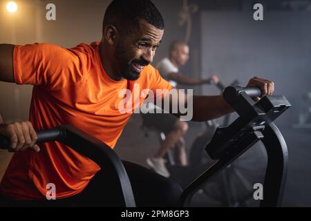 Engagierter birassistischer junger Mann, der im Fitnessstudio mit dem Fahrrad fährt Stockfoto