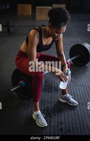 Blick von oben auf müde, birassische junge Frau mit Wasserflasche, die auf der Barbell im Fitnessraum sitzt Stockfoto