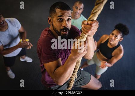 Blick aus einem hohen Winkel auf birassische Freunde, die jubeln und einen jungen Mann anschauen, der im Fitnessstudio auf ein Seil klettert Stockfoto
