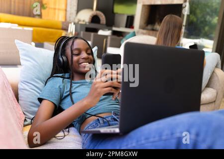 Glückliche, vielfältige Teenager-Freundinnen, die auf dem Sofa sitzen und Smartphones und Laptops verwenden Stockfoto