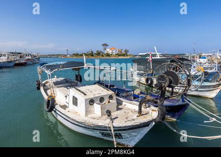 Ein Fischerboot mit einer großen Netzwinde am Bug liegt vor Anker im Fischereihafen von Famagusta, Türkische Republik Nordzypern (TRNK), eine türkisch-zypriotische Flagge weht vom Bug eines benachbarten Bootes. Die weiße Villa im britischen Kolonialstil liegt auf einer kleinen Landzunge im Hintergrund. Stockfoto