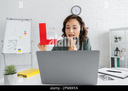 Sprachtherapeut im Headset hält während des Online-Unterrichts Brief und zeigt auf den Mund auf einem Laptop im Sprechzimmer, Stockbild Stockfoto