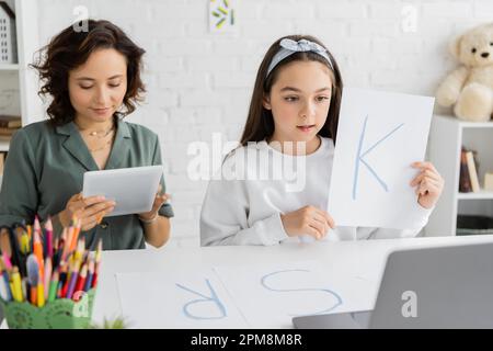 Ein junges Mädchen hält Papier mit Buchstabe K während der Sprachtherapie-Onlinestunde auf einem Laptop in der Nähe der mutter zu Hause, Stockbild Stockfoto