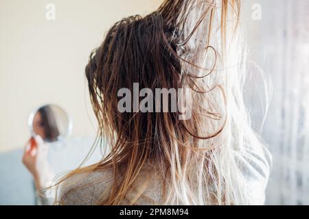 Nahaufnahme einer Frau, die schmutziges öliges und fettiges Haar berührt und zu Hause in den Spiegel schaut. Zeit, den Kopf mit hochwertigem Shampoo zu waschen. Rückansicht Stockfoto