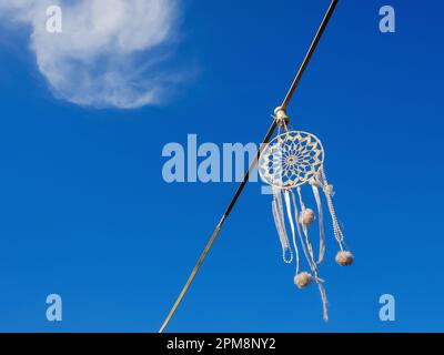 Handgefertigter weißer Traumfänger, der an einer Lichterkette am blauen Himmel hängt, Dekoration der Sommerterrasse des Cafés Stockfoto