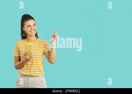 Attraktive junge Frau, die ein Handy in einer Hand hält, mit ihren Zeigefingern zur Seite zeigt, wirbt für einen Platz für ihre Werbeinhalte Stockfoto