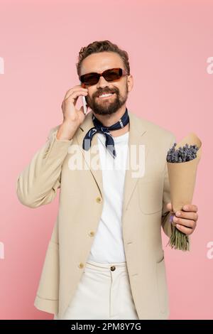 Glücklicher Mann in stilvoller Sonnenbrille und Halstuch mit Blumen und Gespräch auf Smartphone isoliert auf pinkfarbenem Stock-Bild Stockfoto