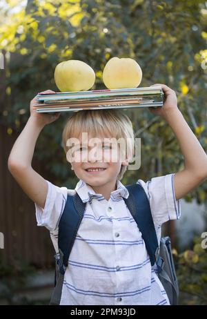 Ein süßer, fröhlicher 6-jähriger Junge mit einem Stapel Bücher und zwei großen Äpfeln auf dem Kopf, der sich für die Schule fertig macht. Bücher lesen, Interessantes Kindermädchen Stockfoto