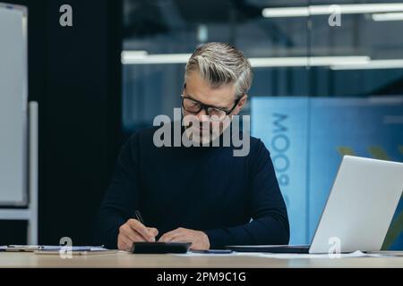 Ein älterer Geschäftsmann sitzt im Büro an einem Tisch mit einem Laptop und schreibt Dokumente. Arbeiten an einem Projekt, Termin, Überstunden, nachts. Stockfoto