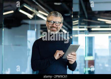 Porträt eines gut aussehenden, grauhaarigen Geschäftsmanns mit Brille, der im Büro stand, ein Tablet benutzte und mit einem Lächeln in die Kamera schaute. Stockfoto