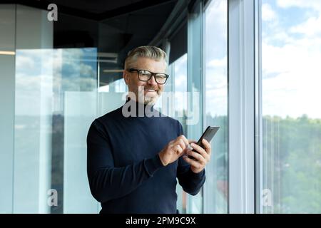 Porträt eines gutaussehenden, grauhaarigen Geschäftsmanns in Brille, der in einem Wolkenkratzer-Büro neben dem Fenster steht, ein Handy hält und benutzt, in die Kamera schaut und lächelt. Stockfoto