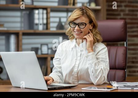Porträt einer erfahrenen, schönen Anwältin, Notar, der im Büro am Tisch sitzt, an einem Laptop und mit Dokumenten arbeitet. Lächelnd und selbstbewusst in die Kamera. Stockfoto