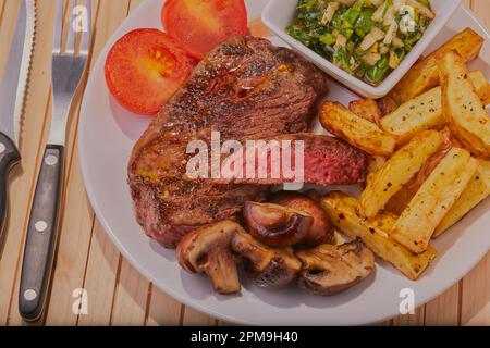 Mittelseltenes Rumpsteak mit Chips, Pilzen und Tomaten mit Chimichurri-Sauce. Stockfoto