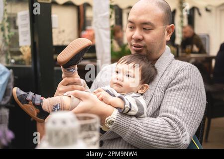 Flexibler, männlicher Säugling, der auf den Beinen seines Vaters sitzt, seine eigenen Beine hält, mit dem Vater spielt, ernster Ausdruck, im Biergarten in einem Pub in Schottland. Happ Stockfoto