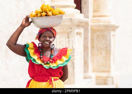 Fröhlicher, lächelnder Palenquera-Straßenverkäufer mit frischem Obst in der Altstadt von Cartagena, Kolumbien. Fröhliche afrokolumbianische Frau in traditionellen Kostümen. Stockfoto