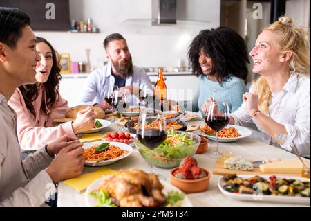 Fröhliche Gruppe von Freunden, die Spaghetti-Pasta mit roter Tomatensoße zu Hause essen, fröhliche junge Leute, die gemeinsam Mittagspause machen Stockfoto
