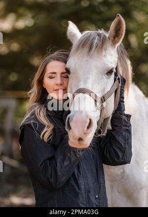 Junge Frau lehnt sich an weißes arabisches Pferd, Augen geschlossen, unscharfer Baumhintergrund, Nahaufnahme Stockfoto