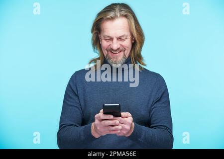 Ein erwachsener Mann liest Nachrichten auf seinem Smartphone und lächelt vor blauem Hintergrund Stockfoto