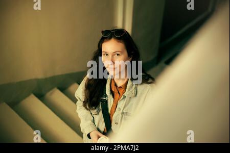 Porträt einer wunderschönen jungen Studentin, die mit einer Tasse Kaffee die Treppe hochklettert. Nach einer Pause zurückkehren. Geschäftsfrau. Stockfoto