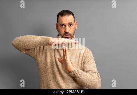 Bärtiger hispanischer Mann in seinen 40s Jahren mit beigefarbenem Rollkragenpullover, der die Auszeit-Geste machte, während er ernsthaft in die Kamera schaute, isoliert im grauen Studio Stockfoto