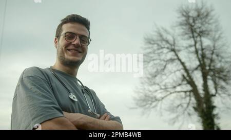 Lächelnder junger großer Doktor mit gekreuzten Armen und Stethoskop, der auf die Kamera schaut Stockfoto