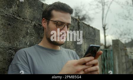 Junger Mann mit Brille, der mit Smartphone Outdoor Spaß in sozialen Medien hat Stockfoto