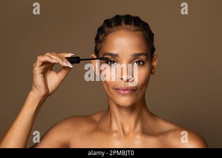 Wunderschöne afroamerikanische Frau mittleren Alters, die Wimpern mit Wimpern mit isolierter Bürste auf braunem Hintergrund mit Mascara bedeckt Stockfoto