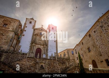 Blick auf die barocke Kirche San Francisco Javier de Caceres, Spanien, auf der Plaza de San Jorge bei Sonnenuntergang mit der Sonne im Hintergrund und Tauben am Himmel Stockfoto