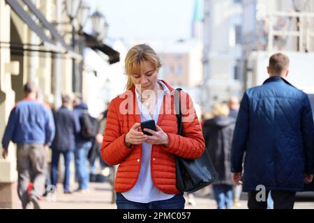 Frau in roter Jacke, die eine Straße entlang läuft, mit Smartphone im Hintergrund. Handy-Nutzung in der Frühlingsstadt Stockfoto