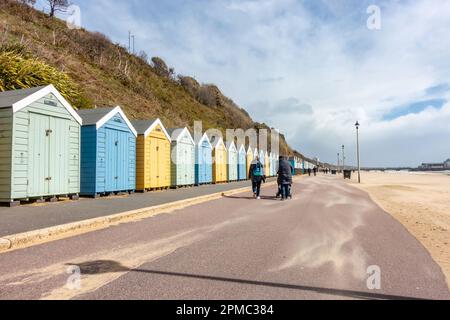 Die Menschen gehen einen Pfad vor den Strandhütten am Bournemouth Beach in Dorset, Großbritannien, unter stürmischem, grauem Himmel mit Sand, der im Wind weht Stockfoto