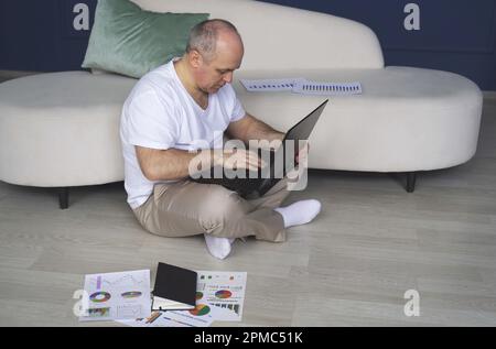 Ein Mann sitzt auf dem Boden neben dem Sofa und arbeitet an einem Laptop. Stockfoto