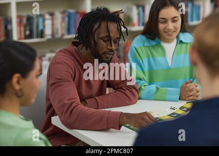 Porträt eines jungen schwarzen Mannes, der Brettspiele mit einer Gruppe von Freunden spielt und ein bewegliches Spielstück Stockfoto