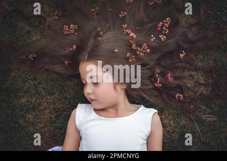 Kleines Mädchen in weißem Kleid, das auf dem Gras liegt, schläft, ihr Haar ist sehr lang und prinzessenartig, sie spielt mit ihren Händen am Thema „Kiefer“ Stockfoto