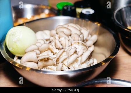 Austernpilze in einer Metallschüssel auf einem Holztisch mit geschälten Zwiebeln. Rohe Zutaten vor dem Kochen. Stockfoto