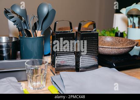 Küchenutensilien und Werkzeuge wie Spatel oder Schneebesen, graue Schürze und zwei handgehaltene Stahlgräber stehen vor dem Kochkurs auf einem Tisch. Kulinarischer Meister cla Stockfoto