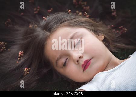 Kleines Mädchen in weißem Kleid, das auf dem Gras liegt, schläft, ihr Haar ist sehr lang und prinzessenartig, sie spielt mit ihren Händen am Thema „Kiefer“ Stockfoto