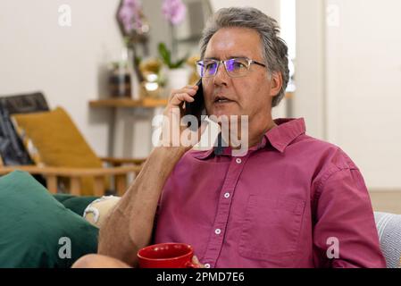Ein weißer Senior-Mann hält eine Kaffeetasse und spricht über ein Smartphone, während er auf dem Sofa sitzt Stockfoto
